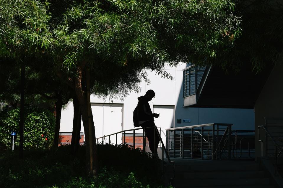 A student stands at East Los Angeles College under a tree looking at his phone.